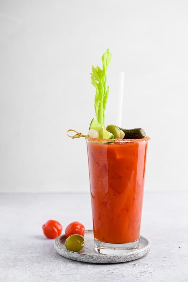 Tomato-red Bloody Mary in a tall glass with celery, citrus, savory garnish, and a straw on a pale surface