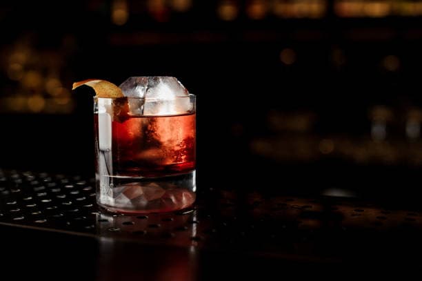 Amber-red Boulevardier in a rocks glass with a large clear ice cube and orange peel garnish on a dark bar surface