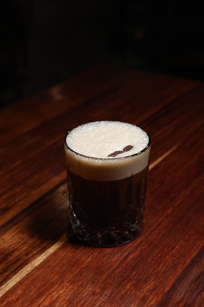Dark Carajillo in a cut-glass tumbler with a thick foamy top and two coffee beans on a wooden surface