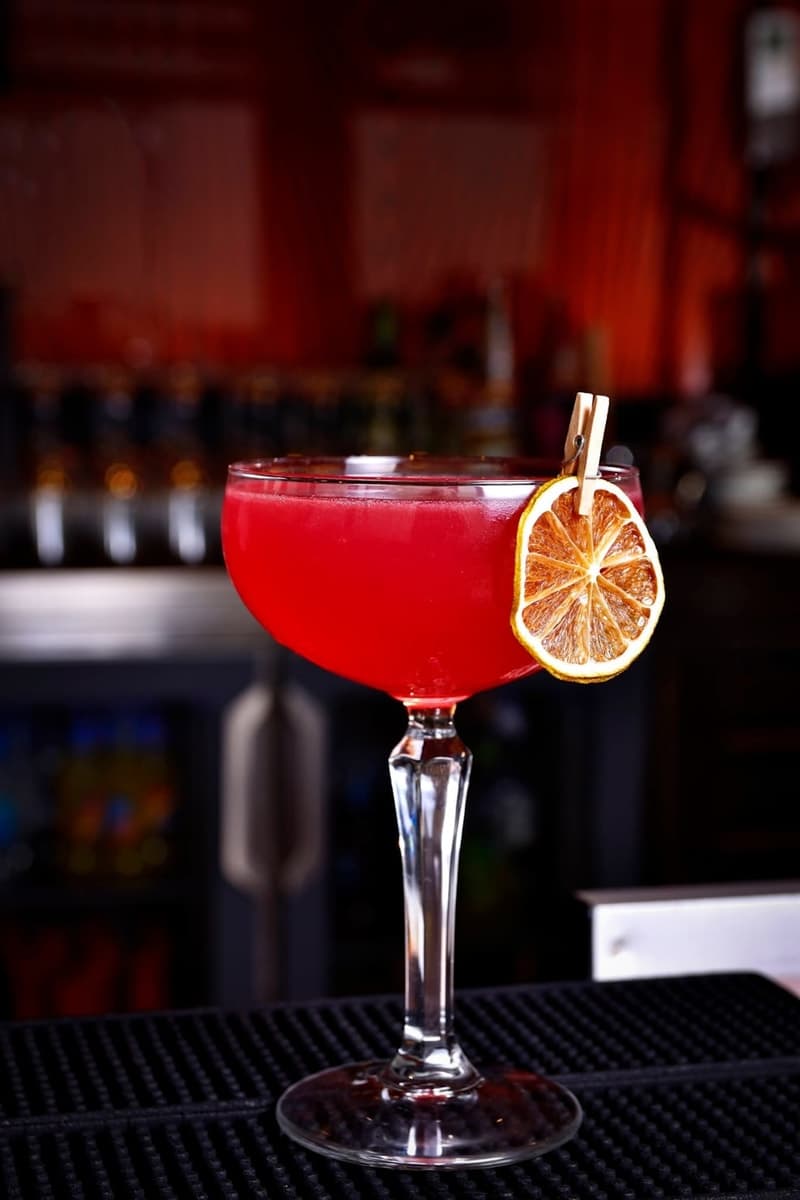 Red-pink Cosmopolitan in a stemmed cocktail glass with a dried citrus wheel garnish on a bar mat
