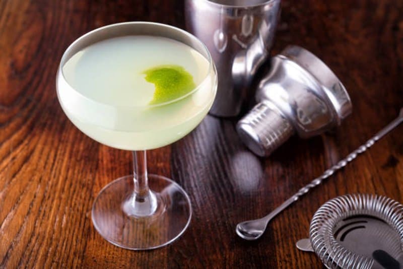 Pale Daiquiri in a coupe with a lime twist beside metal bar tools on a wooden bar