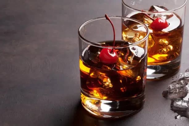 Two amber Manhattan-style cocktails in rocks glasses with red cherry garnishes on a dark tabletop
