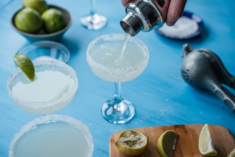 Salt-rimmed Margaritas on a blue table with lime wedges and a shaker pour