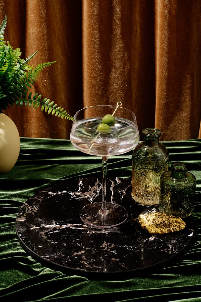 Clear Martini in a coupe glass with olives on a pick against velvet and glass bottles