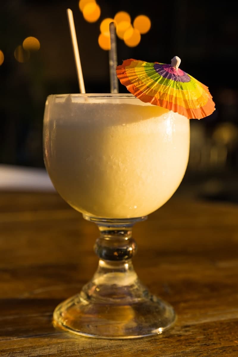 Creamy pale-yellow Piña Colada-style cocktail in a stemmed goblet with two straws and a colorful paper umbrella