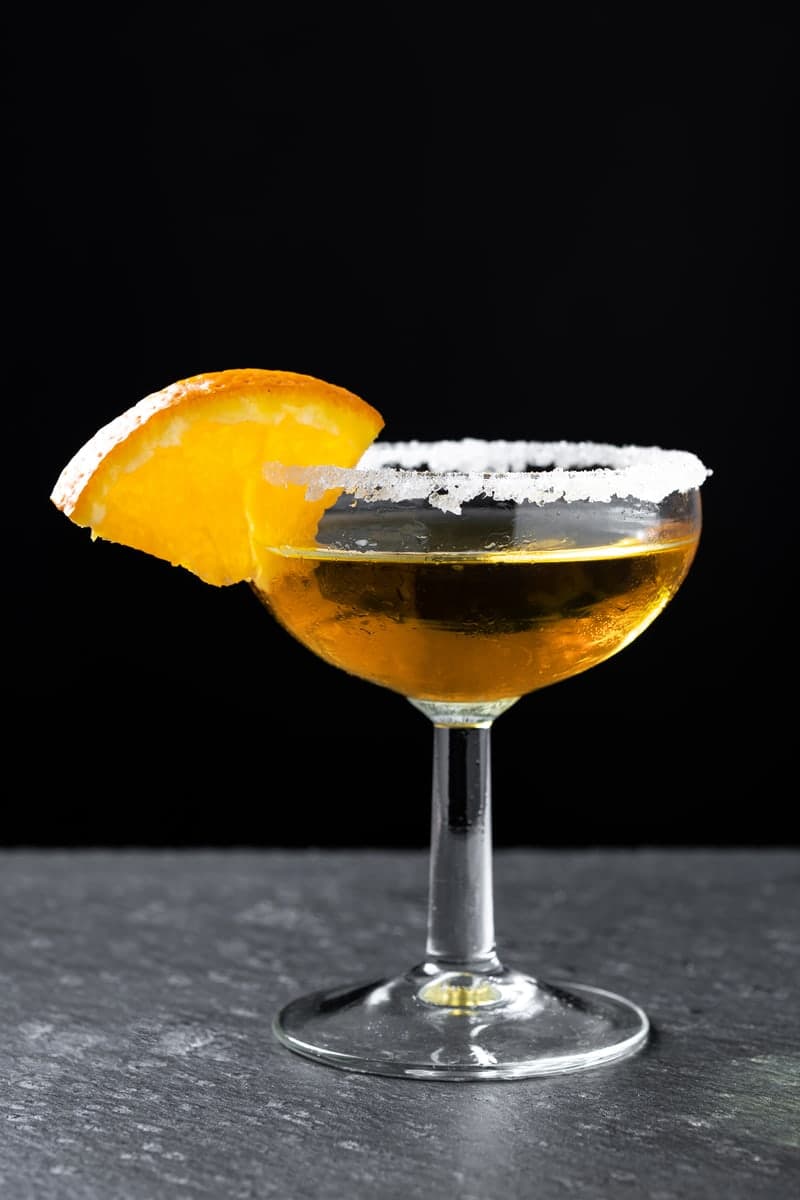 Golden Sidecar-style cocktail in a sugar-rimmed coupe with an orange wedge on a gray wooden tabletop against a black background