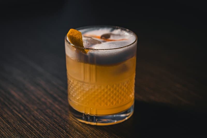Foamy Whiskey Sour in a rocks glass on dark wood with lemon peel and bitters on top